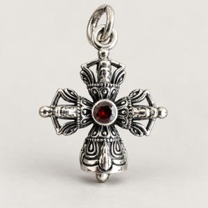 Op de afbeelding: Een zilverkleurige metalen hanger in de vorm van een gekruiste vajra, met gedetailleerde reliëfversieringen en een ronde, donkerrode gefacetteerde edelsteen in het midden. De hanger is voorzien van een stevige, ronde ring aan de bovenzijde.