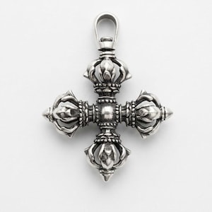 Könnte beinhalten: Ein silberfarbener Metallanhänger in Form eines Doppel-Vajra, auch bekannt als Vishvavajra. Das Objekt besteht aus vier symmetrischen Zacken, die von einer zentralen Kugel ausgehen, mit detaillierten Gravuren und einer polierten Oberfläche, versehen mit einer Öse.