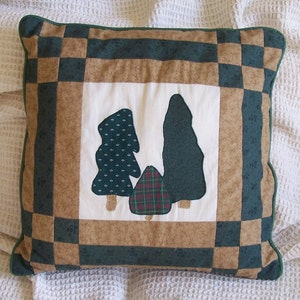May include: Square decorative pillow with a patchwork design. The pillow features a central panel with appliqued trees in shades of green and brown. The border has a checkerboard pattern in similar colors, with a green trim.