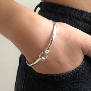 May include: A polished silver bangle bracelet featuring a circular hook-and-eye style clasp mechanism. The bracelet has a smooth, rounded band that forms a continuous loop, with the two ends meeting at the prominent, circular interlocking closure.