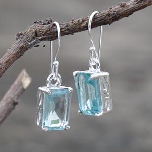 May include: A pair of drop earrings featuring rectangular, emerald-cut light blue gemstones set in silver metal frames. Each earring hangs from a simple silver hook, showcasing a clear, faceted stone design with visible metal prongs and side detailing.