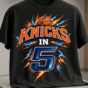 Op de afbeelding: Een zwart t-shirt met een grafisch ontwerp met de oranje tekst KNICKS IN en een groot blauw cijfer 5. Het ontwerp bevat een basketbal, oranje en blauwe bliksemschichten en explosieve energie-effecten rondom de centrale tekst en het cijfer.