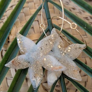 Peut inclure: Une paire de boucles d'oreilles pendantes en forme d'étoile à cinq branches en résine transparente. La résine est incrustée de sable beige, de paillettes dorées et de petits coquillages blancs. Chaque étoile est suspendue à un anneau en métal doré.