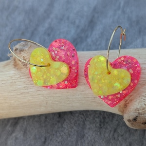 Peut inclure: Une paire de boucles d'oreilles créoles avec deux breloques en forme de cœur superposées. Le grand cœur est rose vif avec des paillettes, tandis que le petit cœur est jaune néon avec des paillettes épaisses. Les cœurs pendent à des anneaux dorés.