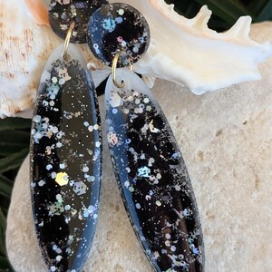 Peut inclure: Une paire de boucles d'oreilles pendantes composée d'une pièce supérieure circulaire et d'une goutte ovale allongée, en résine noire incrustée de paillettes hexagonales et fines multicolores, reliées par des anneaux en métal doré.