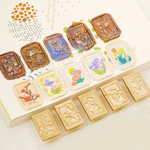 May include: A collection of rectangular wax seals and matching metal stamps featuring floral and botanical designs. The seals display various colors including brown, blue, and white, with some painted in vibrant hues, while the stamps are gold-toned.