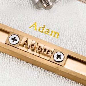 Könnte beinhalten: Ein Prägewerkzeug aus Messing mit einer horizontalen Schiene und einzelnen Buchstabenblöcken, die das Wort Adam bilden, befestigt durch zwei silberfarbene Kreuzschlitzschrauben. Das Werkzeug liegt auf einer weißen, strukturierten Oberfläche mit goldener Prägung.