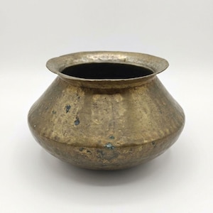 May include: A small, round, hammered brass pot with a wide, flared rim. The metal surface features a weathered, aged patina with visible dark spots and a textured, uneven finish throughout the exterior body of the vessel.