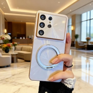 Puede incluir: Una funda de smartphone de color beige claro con un módulo de cámara rectangular que incluye cuatro lentes circulares y un flash central. La parte posterior de la funda presenta un anillo de soporte plateado centrado en la superficie lisa.