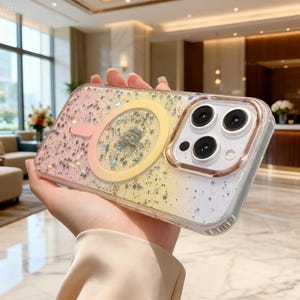 May include: A clear smartphone case featuring a gradient design that transitions from pink to yellow and white, embedded with silver glitter flakes. The case includes a circular magnetic charging ring and a metallic rose gold camera lens frame.