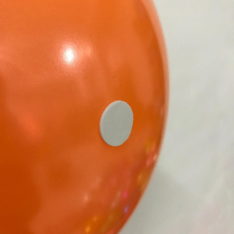 May include: A smooth, vibrant orange surface featuring a single, small, circular white sticker centered on the curved area. The material appears glossy and reflective, with a bright orange hue dominating the frame and a clean white circular accent.