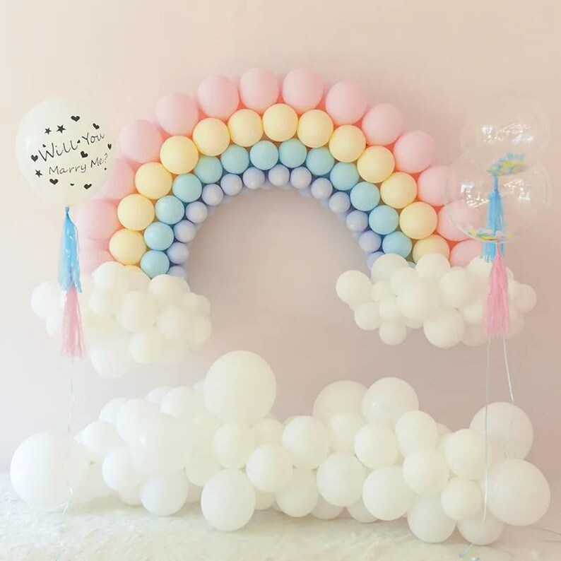 May include: A balloon arch shaped like a rainbow with pink, yellow, and blue balloons, anchored by white balloon clouds. A single white balloon on the left features the black text Will You Marry Me? with small heart icons and a pink tassel.