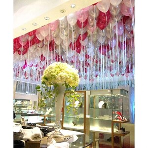 May include: A tall, clear glass trumpet vase filled with a large, rounded bouquet of white hydrangea flowers. Above the vase, the ceiling is densely covered with a multitude of floating white, pink, and magenta balloons with long white ribbons.