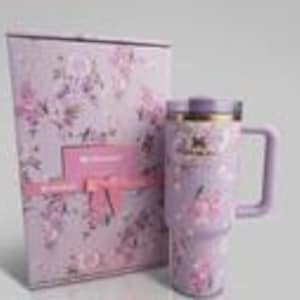 May include: A light purple stainless steel tumbler with a floral pattern, a matching lid, and a side handle, displayed next to a rectangular gift box featuring the same lavender floral print, a pink ribbon accent, and the Stanley brand logo.