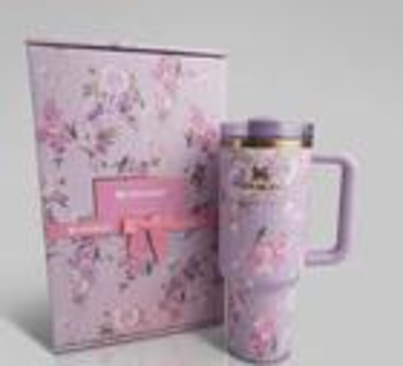 May include: A light purple stainless steel tumbler with a floral pattern, a matching lid, and a side handle, displayed next to a rectangular gift box featuring the same lavender floral print, a pink ribbon accent, and the Stanley brand logo.