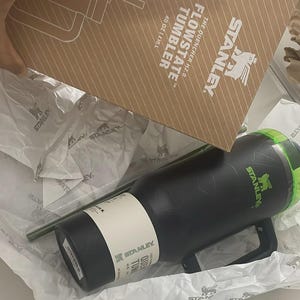 May include: A black Stanley Flowstate Quencher H2.0 tumbler with a green lid and handle, featuring a white product label. The tumbler is packaged in a brown cardboard box printed with the Stanley logo and the text 40 OZ | 1.18 L.