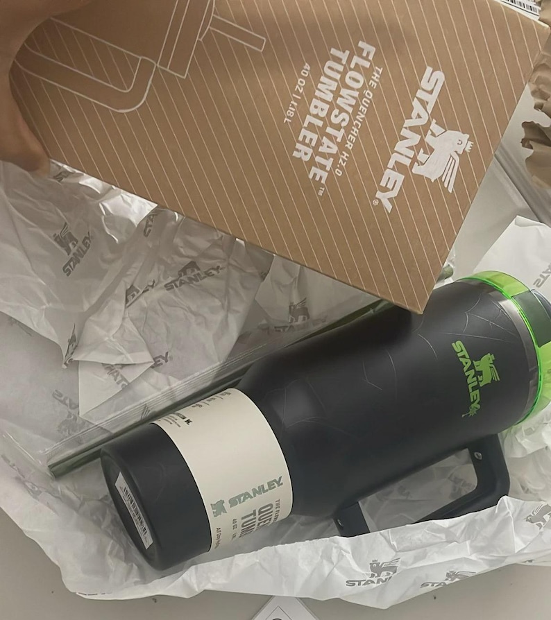 May include: A black Stanley Flowstate Quencher H2.0 tumbler with a green lid and handle, featuring a white product label. The tumbler is packaged in a brown cardboard box printed with the Stanley logo and the text 40 OZ | 1.18 L.