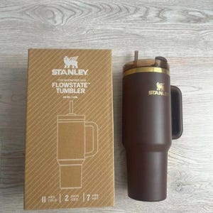 May include: A dark brown Stanley Flowstate tumbler with a handle and a straw, next to its cardboard packaging. The box features the Stanley logo, the text "The Quencher H2.0 Flowstate Tumbler 40 oz / 1.18L", and icons indicating 11 hours cold, 2 days iced, and 7 hours hot.