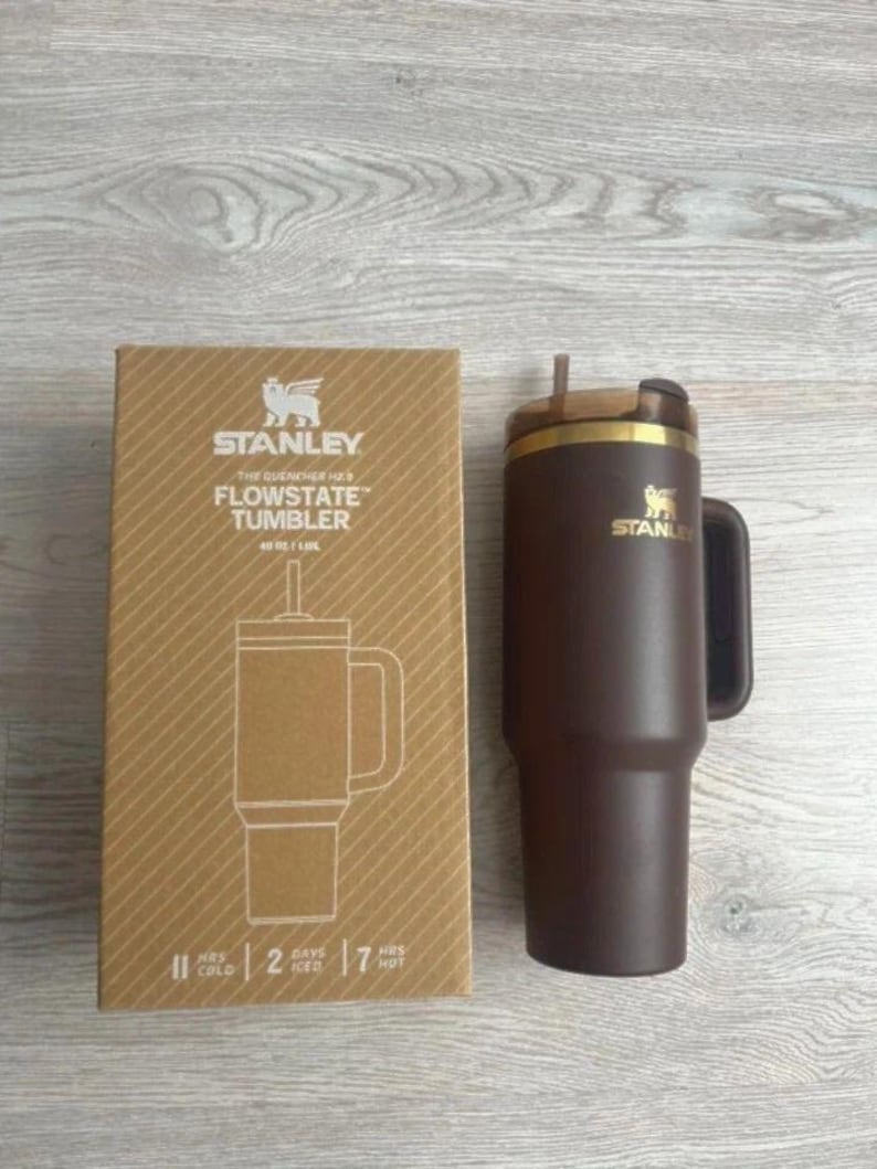 May include: A dark brown Stanley Flowstate tumbler with a handle and a straw, next to its cardboard packaging. The box features the Stanley logo, the text "The Quencher H2.0 Flowstate Tumbler 40 oz / 1.18L", and icons indicating 11 hours cold, 2 days iced, and 7 hours hot.