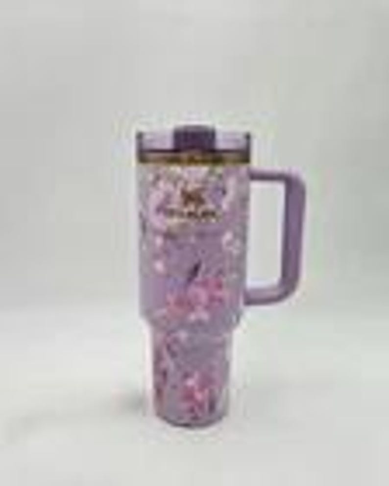 May include: A light purple insulated tumbler with a floral pattern featuring pink and white blossoms. The tumbler includes a dark purple lid, a sturdy side handle, and the Stanley brand logo printed in the center of the upper section.