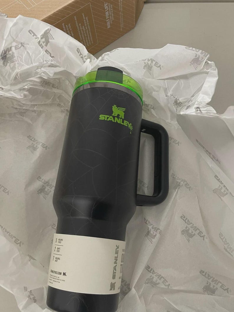 May include: A black Stanley tumbler with a spiderweb pattern, a lime green lid, and a black handle. The tumbler features a white paper band around the base with text indicating 11 hours cold, 2 days iced, and 7 hours hot, along with the Stanley logo.