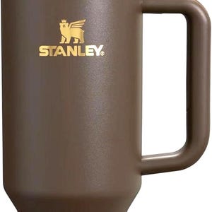 May include: A matte brown insulated tumbler with a large side handle, a matching brown straw, and a dark translucent lid. The upper portion features a gold-coloured band and the Stanley logo with a winged bear emblem printed in gold on the front.