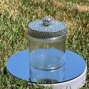 以下が含まれることがあります： ラインストーンで覆われたシルバーの蓋と、小さなラインストーンで覆われたノブが付いた透明なガラス瓶。瓶は鏡面の上にあり、緑の草を反射しています。蓋とノブは日光の下で輝いています。