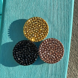 May include: Three round, bedazzled accessories in gold, black, and rose gold. The accessories are covered in small, sparkling crystals. The items are arranged on a textured, turquoise surface.