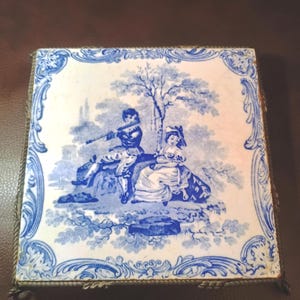 May include: A square ceramic tile featuring a blue and white transferware design of a person playing a flute and another person sitting beside them under a tree. The tile is framed by a decorative blue border and has a metallic rope-style edge.