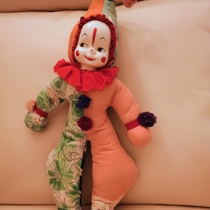 May include: A clown doll with a porcelain face, painted red facial markings, and a pointed hat. The body features a split design with one side in solid peach fabric and the other in a green floral print, accented by a red ruffled collar and pom-poms.