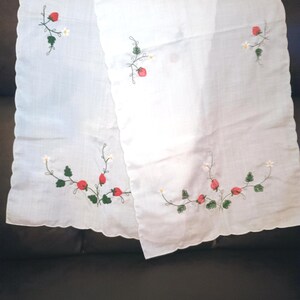 May include: A white rectangular fabric runner with scalloped edges, featuring embroidered red strawberries, green leaves, and small white flowers. A metal ruler is positioned at the bottom, showing a length of approximately 12 inches.
