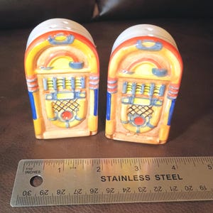 May include: Two ceramic jukebox-shaped salt and pepper shakers featuring yellow, orange, and blue painted details. A stainless steel ruler is placed in front, showing each shaker is approximately 2.5 inches wide at the base.