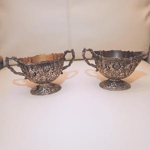 May include: Two silver-toned metal bowls with ornate floral relief patterns, scalloped rims, and pedestal bases. Each bowl features two decorative handles on opposite sides, showcasing intricate embossed sunflower and leaf designs throughout.