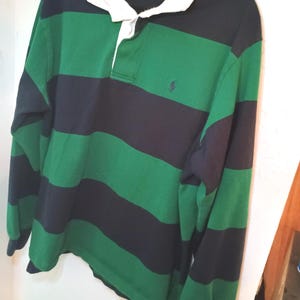 May include: A long-sleeve rugby shirt featuring wide horizontal stripes in dark navy blue and forest green. The shirt has a solid white collar, a buttoned placket, and a small embroidered logo on the left chest. The interior label indicates size XL.