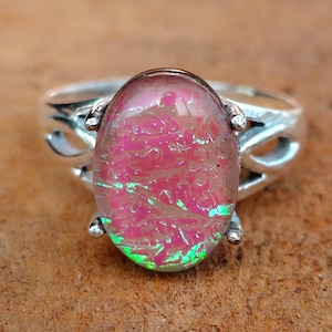 Puede incluir: Un anillo de plata con una piedra cabujón de forma ovalada con un patrón moteado de color rosa vibrante y verde iridiscente. La banda de metal tiene un diseño recortado decorativo en ambos lados, sujetando la piedra central con cuatro garras.
