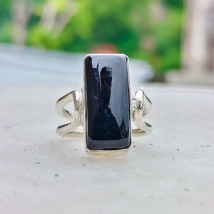 Peut inclure: Une bague en argent ornée d'une pierre noire rectangulaire polie, fixée verticalement au centre. L'anneau est composé de deux fins fils d'argent incurvés qui se rejoignent à la base du sertissage, créant un design ouvert et aéré sur les côtés.
