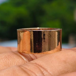 May include: A wide, polished rose gold band ring with a smooth, reflective surface. The ring features a flat, cylindrical profile with clean, straight edges, showcasing a warm metallic hue that catches the light across its entire circumference.