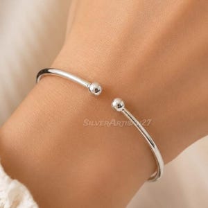 May include: A polished silver cuff bracelet featuring a slender, open-ended band design. The bracelet terminates at both ends with a small, smooth, spherical ball detail. The text SilverArtis27 is visible on the surface of the metal band.