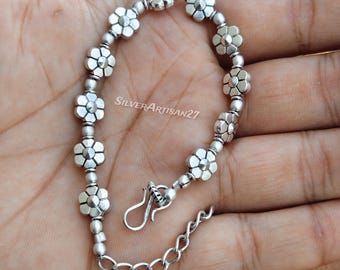 May include: A silver-toned bracelet featuring a series of small, five-petaled flower charms alternating with smooth round beads. The piece includes a hook-style clasp and an adjustable chain link extension. The text SilverArtisan27 is visible.