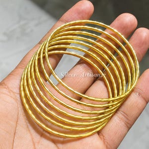 Puede incluir: Un conjunto de siete brazaletes finos de color dorado con una superficie texturizada y martillada. Las pulseras circulares están apiladas juntas, presentando un patrón repetitivo de pequeñas hendiduras a lo largo de toda la circunferencia.