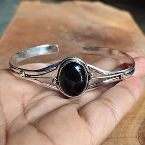 Può includere: Un bracciale rigido in argento con una pietra nera ovale centrale. La fascia metallica si divide in due sottili fili d'argento lucido su ciascun lato della pietra, accentuati da piccole sfere d'argento nei punti di giunzione.