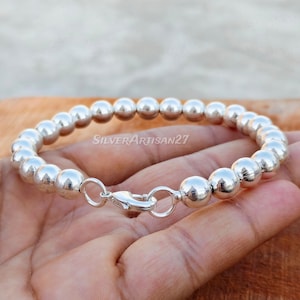 Peut inclure: Un bracelet de perles en argent poli composé d'une série de perles métalliques sphériques et uniformes. Le bracelet est fixé par un fermoir mousqueton en argent et comporte le texte SilverArtisan27 superposé sur l'image.