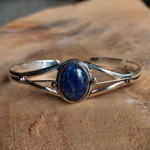 Peut inclure: Un bracelet manchette en argent orné d'une pierre précieuse lapis-lazuli ovale au centre, présentant une couleur bleu profond avec de légères inclusions. La tige métallique se divise en deux fils d'argent polis de chaque côté, finis par des billes.
