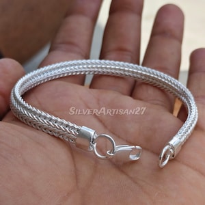 Puede incluir: Una pulsera de cadena plateada con un diseño trenzado de múltiples hilos y acabado pulido. La pulsera incluye una anilla circular y un cierre de mosquetón. El texto SilverArtisan27 es visible en el centro de la imagen.