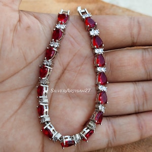 May include: A silver-toned bracelet featuring a series of pear-shaped red gemstones alternating with small, clear, round accent stones. The jewelry piece includes a secure metal clasp closure and displays the text SilverArtisan27 on the surface.