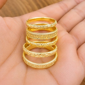 May include: A stack of five gold-toned metal rings, each featuring a distinct engraved pattern. The designs include repeating floral motifs, cross-hatch textures, and beaded edges, all showcasing a polished, reflective finish on the surface.