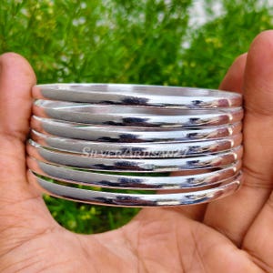Peut inclure: Une pile de sept bracelets joncs argentés polis avec une surface lisse et arrondie. Les bracelets en métal présentent une finition réfléchissante et sont empilés de manière uniforme. Le texte SilverArtisan21 est visible sur la surface.