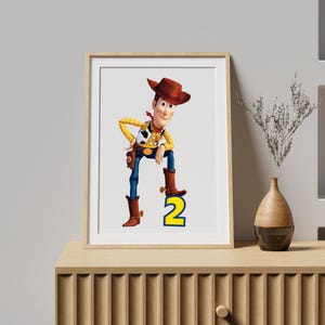 May include: A framed wall art print featuring the character Woody from Toy Story, depicted wearing a brown cowboy hat, yellow shirt, cow-print vest, and blue jeans, with one foot resting on a large, bold, yellow number two with a blue outline.