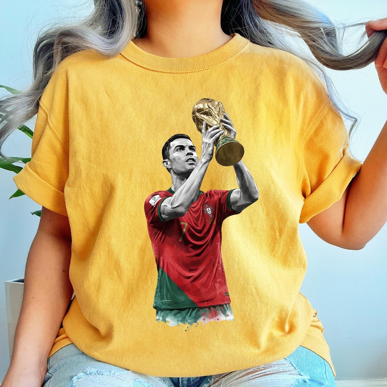 May include: A mustard yellow short-sleeve t-shirt featuring a graphic print of a soccer player in a red and green jersey holding a gold trophy above their head. The player is depicted in a black and white style, while the jersey retains its colors.