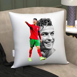 May include: A white square throw pillow featuring a graphic print of a soccer player in a red jersey and green shorts, alongside a large black and white close-up portrait of the same athlete smiling, set against a plain white background.